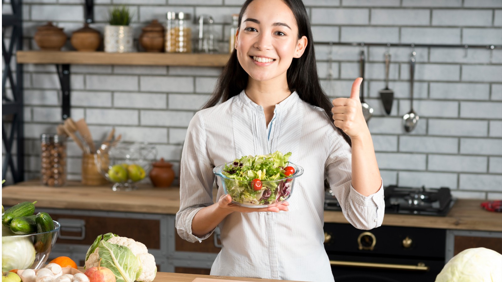 Young woman standing in a kitchen holding a salad and giving a thumbs up